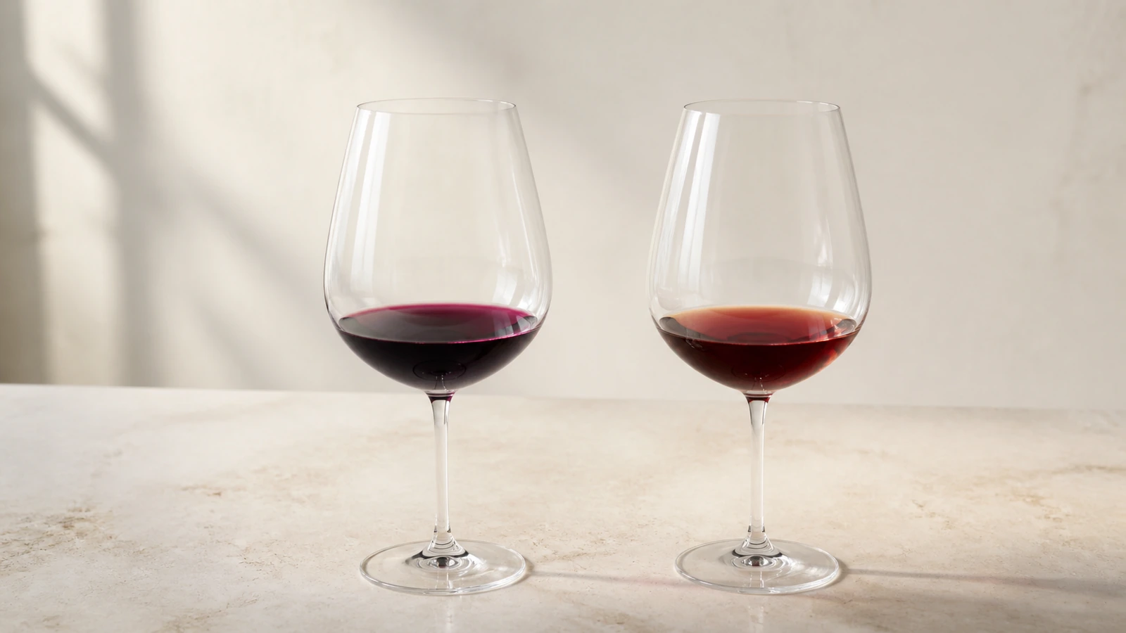 Two clear glasses of red wine on a pale surface, one deep violet-red and the other translucent garnet with a brick-colored rim, illustrating how red-wine color can evolve.